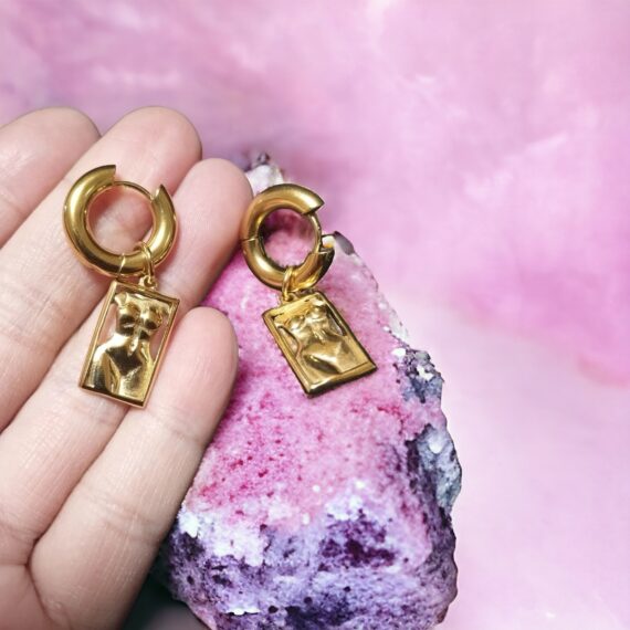 A hand delicately holds "The Body Earrings" against a backdrop of a pink geode, highlighting their exquisite design against the natural beauty of the crystal.
