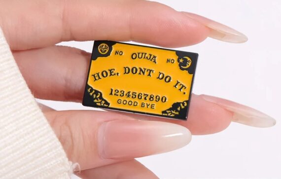 Enamel pin featuring the phrase 'Ouija Hoe, Don't Do It' against a backdrop of a stylized Ouija board design, perfect for adding a touch of occult humor to your attire.