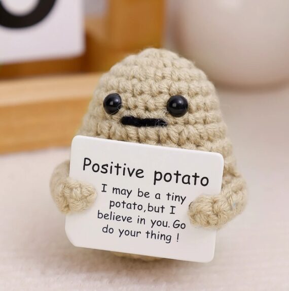 A close-up of the Positive Potato Doll, showcasing its hand-knitted texture and adorable facial features. The doll holds a tiny sign that reads, "Positive Potato: I might be a tiny potato, but I believe in you. Go do your thing!